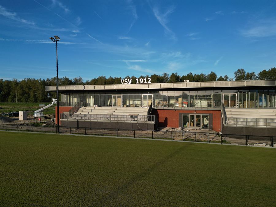 Clubhuis met de naam VSV 1912 op de gevel en een lichtmast tegen een blauwe lucht