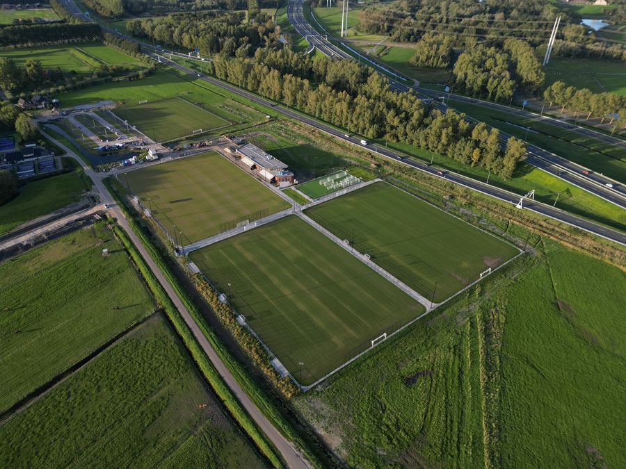 Velden en clubhuis met de bomenrij en de weg langs het park